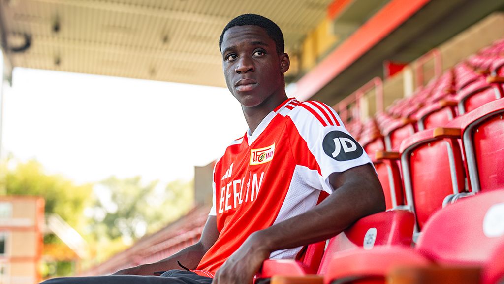 A soccer player sits in an empty stadium, wearing a red jersey with white details, and looks pensively into the distance.