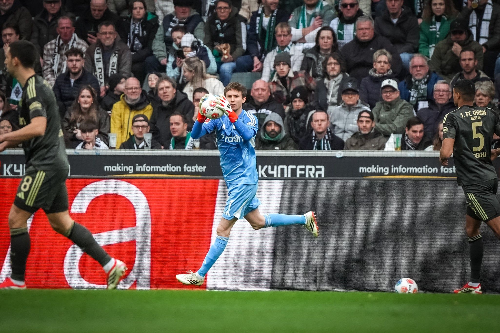 Torwart in blauer Ausrüstung fängt den Ball, während Spieler und Fans im Hintergrund zu sehen sind. Stadionatmosphäre und Spielfeld sichtbar.