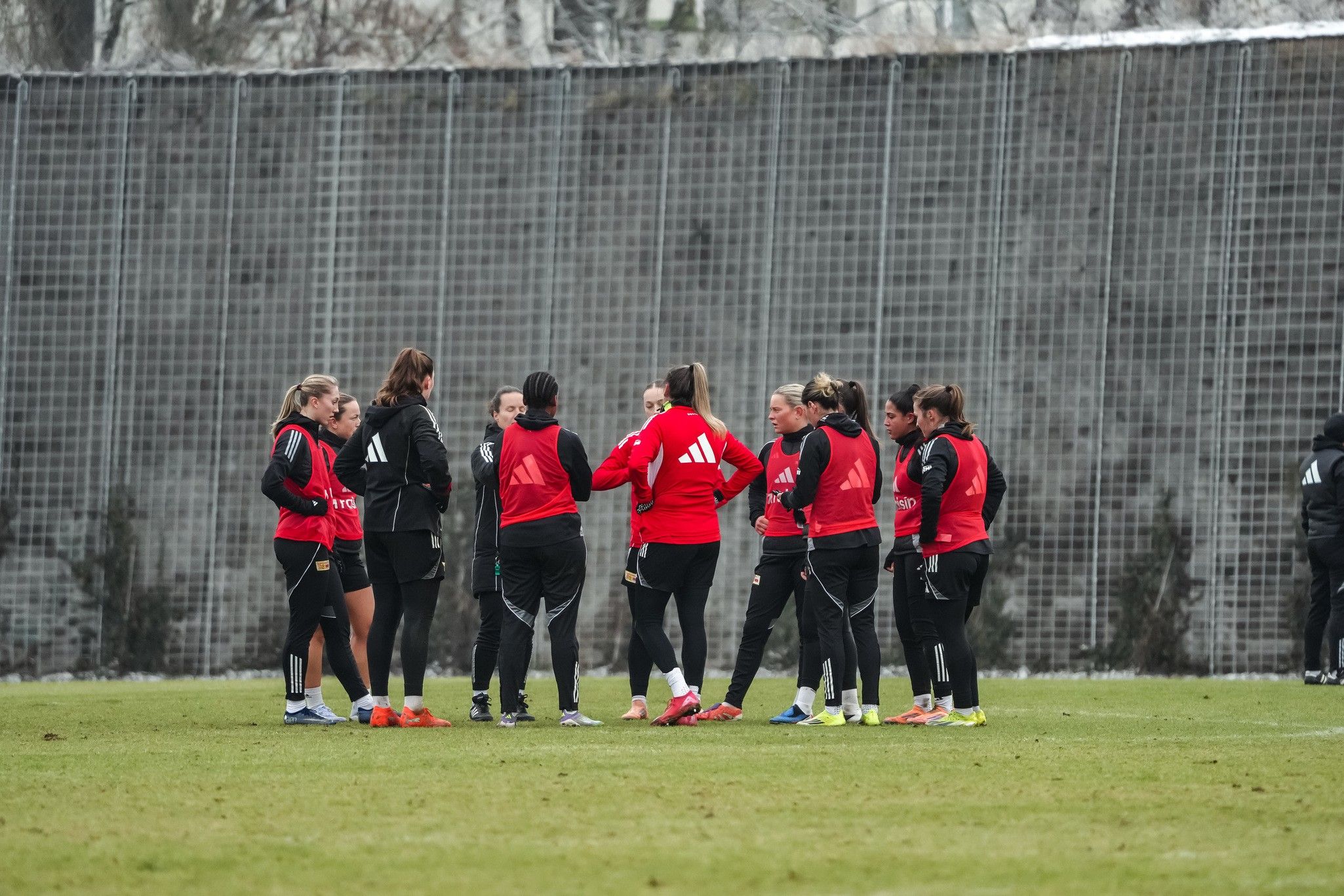 Eine Gruppe von Fußballspielerinnen in roten Trainingsjacken steht auf einem Trainingsplatz und bespricht sich.