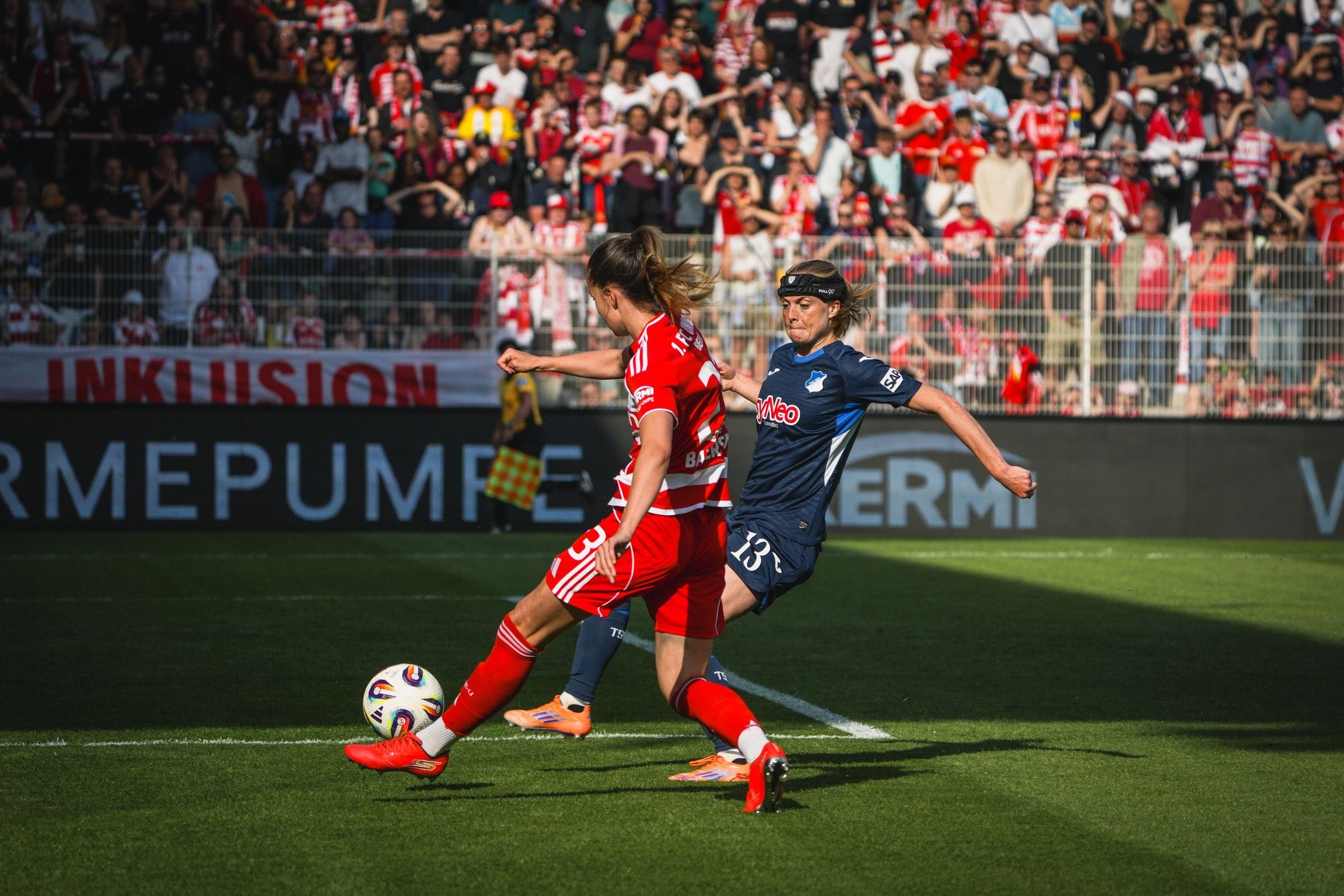 Eine Fußballspielerin in rotem Trikot schießt den Ball, während eine Gegnerin im blauen Trikot versucht, sie zu verteidigen.