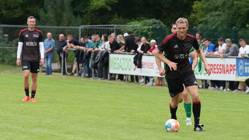 Zwei Fußballspieler stehen auf einem Sportplatz, während eine Menge im Hintergrund zuschaut. Beides trägt sportliche Kleidung.