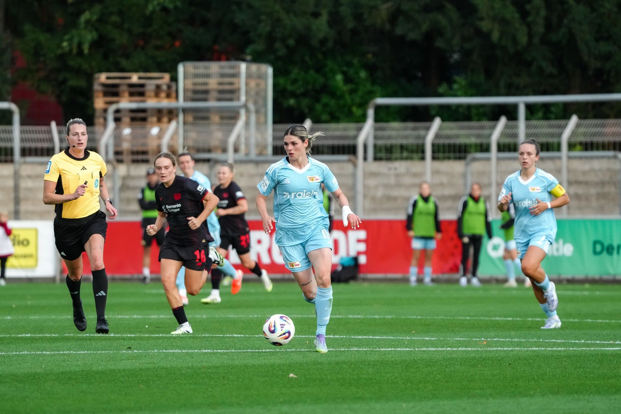 A female soccer player in a light blue jersey sprints across the field while her teammates and opponents stand further away.