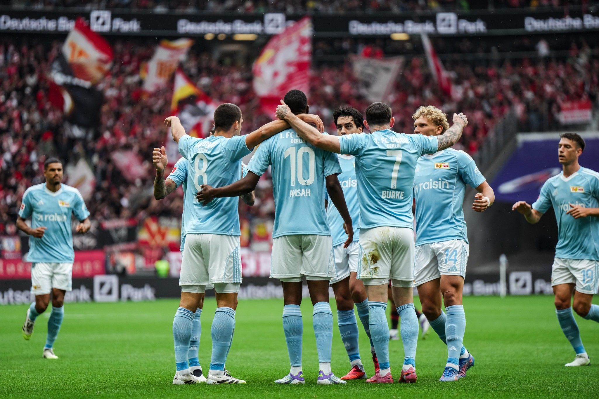 Fußballmannschaft in hellblauen Trikots feiert ein Tor vor jubelnden Fans im Hintergrund.