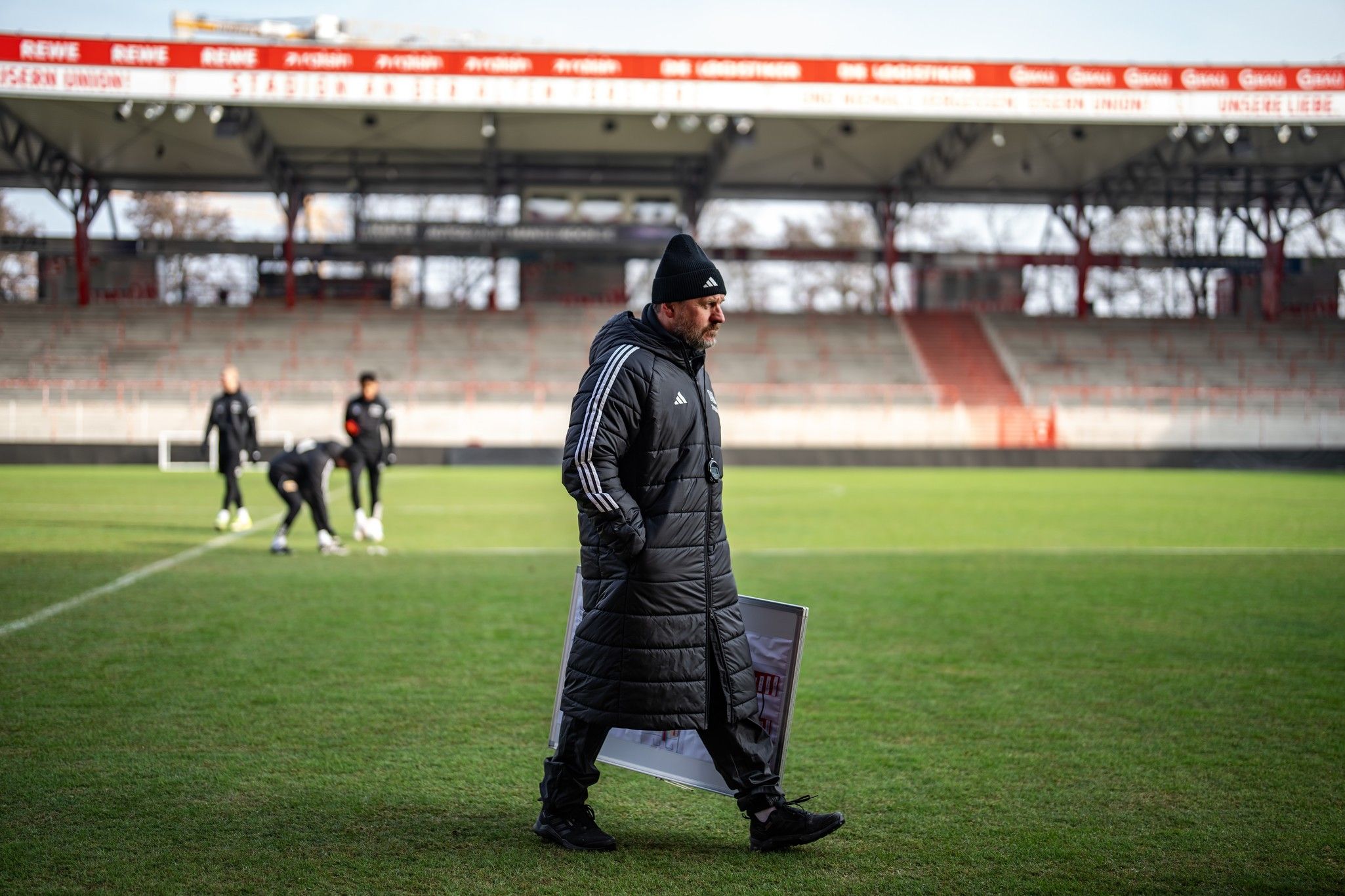 Ein Trainer in einer langen Jacke mit einer Mütze geht auf einem Fußballfeld, während Spieler im Hintergrund trainieren.