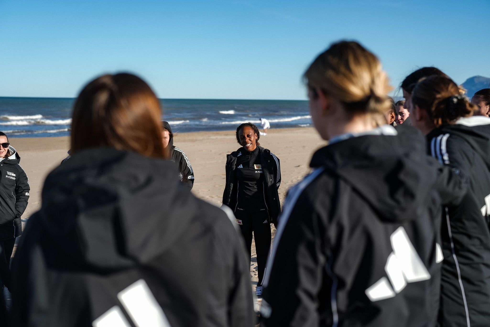 Gruppentraining am Strand; Frauen in dunkler Sportbekleidung hören einer Trainerin zu. Klarer Himmel und Meer im Hintergrund.