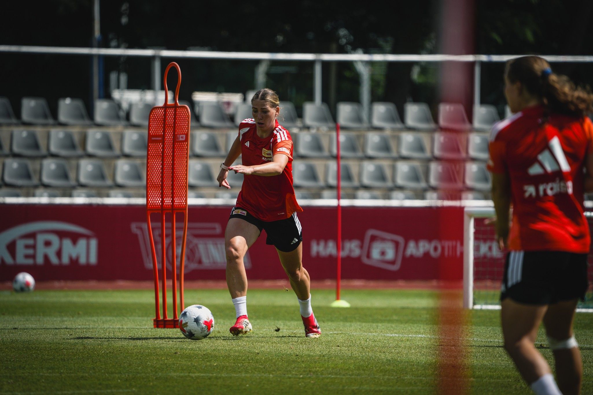 Eine Fußballspielerin in rotem Trikot übt dribbeln mit einem Trainingsdummy auf dem Platz. Im Hintergrund sind leere Sitzreihen zu sehen.