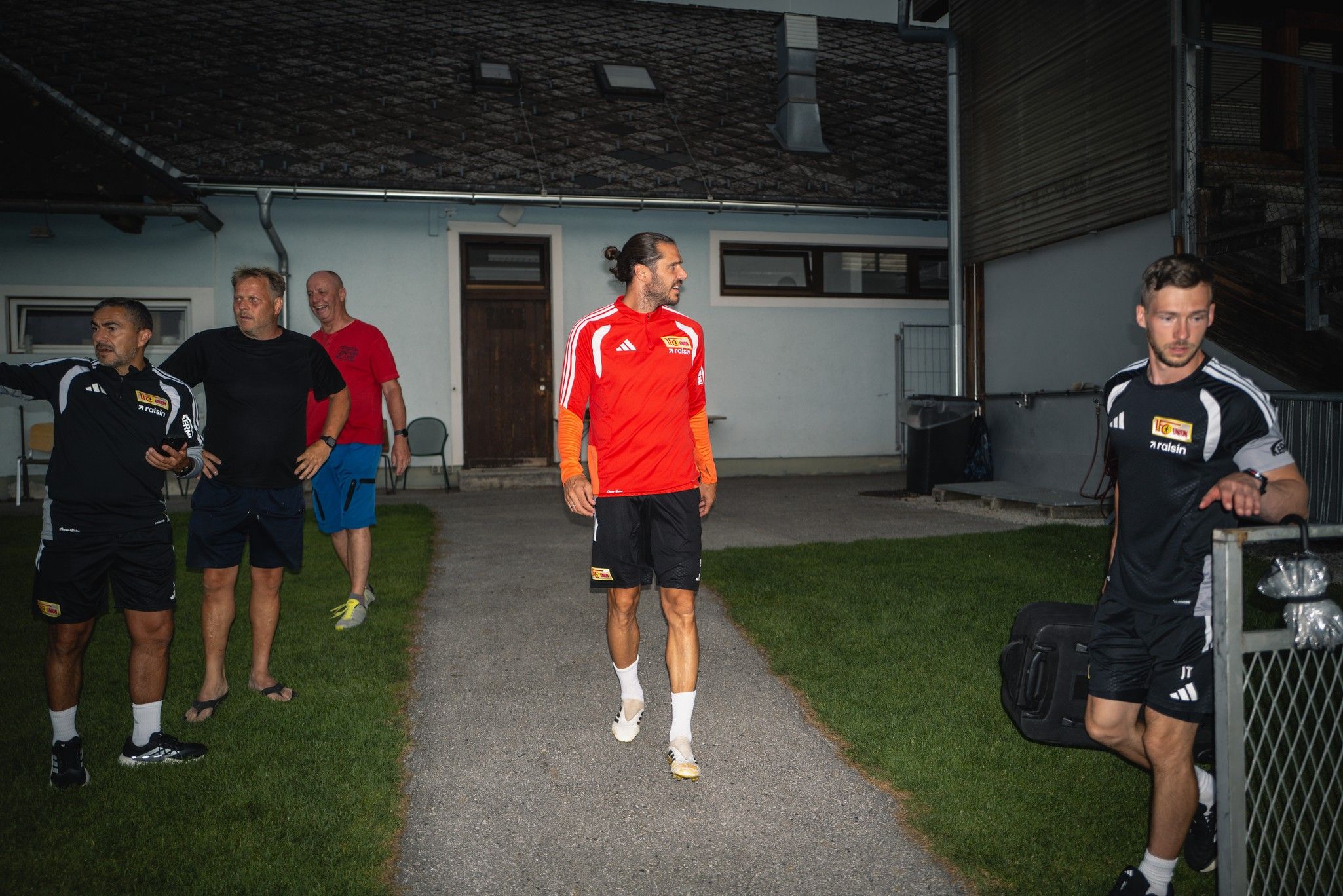 Vier Männer in Sportkleidung, einer mit einem roten Trikot, gehen auf einem Weg. Im Hintergrund ist ein Gebäude zu sehen.
