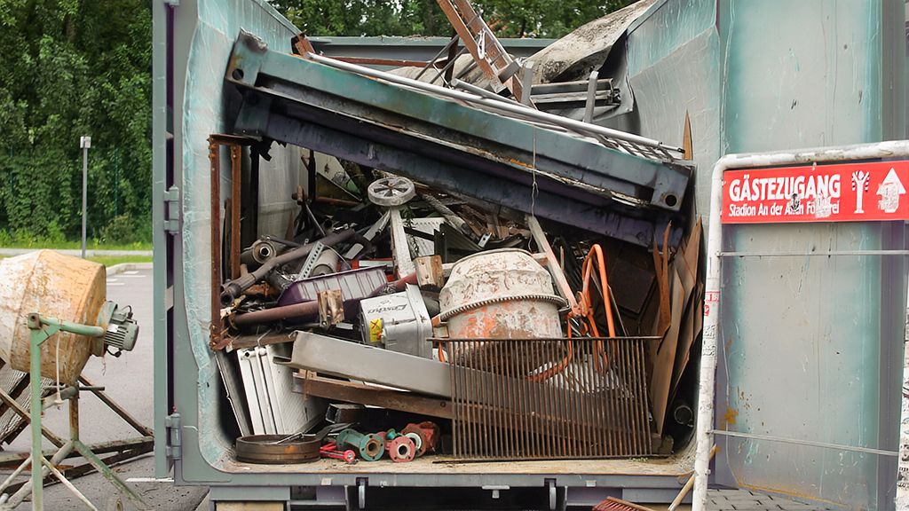 Ein überfüllter Container mit einer Vielzahl von Schrott und Abfällen, darunter Metallteile und alte Geräte.
