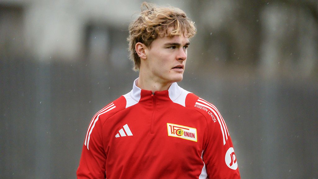A young soccer player in a red tracksuit with white accents stands thoughtfully on a training field.