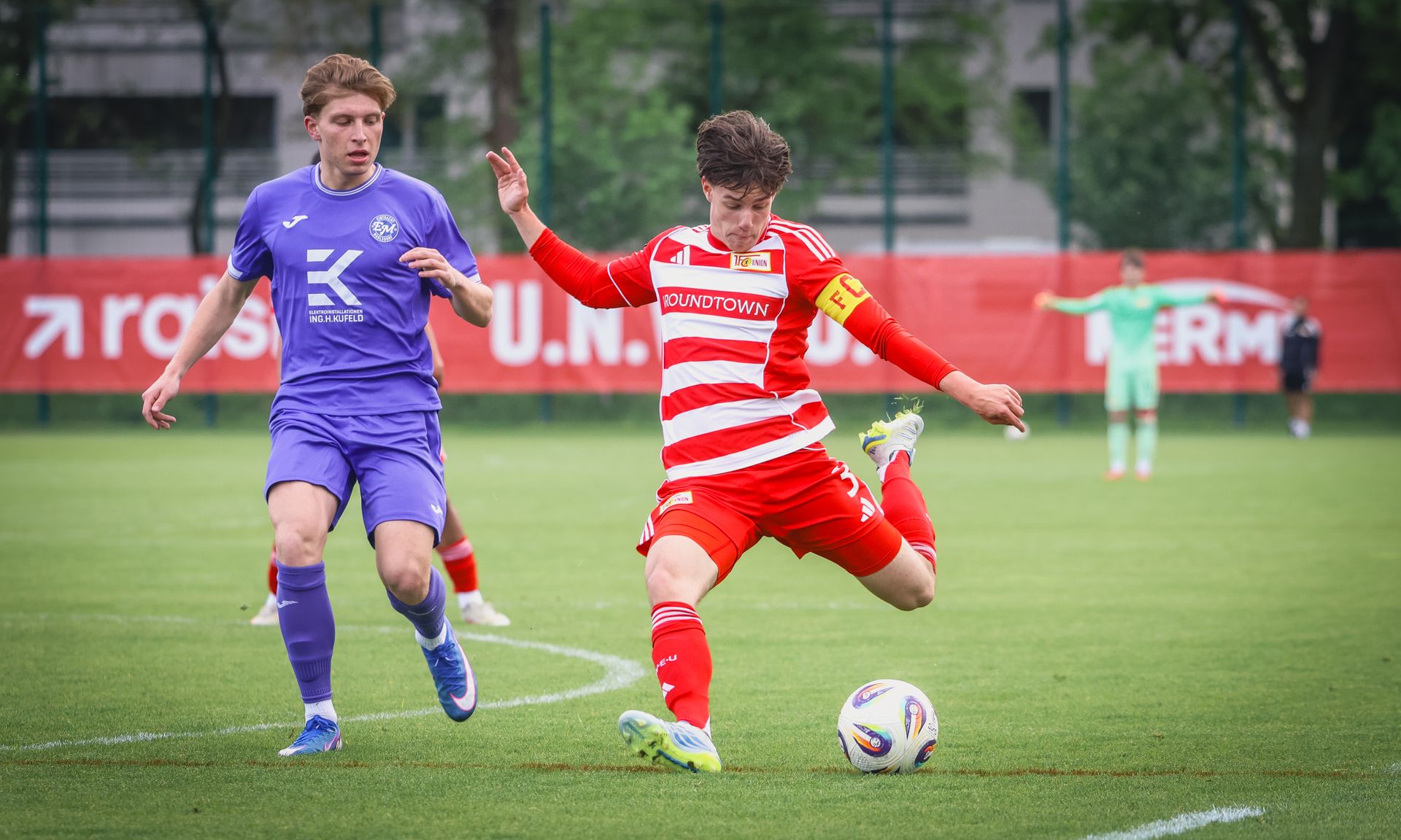 Ein junger Fußballspieler in rotem Trikot schießt den Ball, während ein Gegenspieler in lila ihn verfolgt.