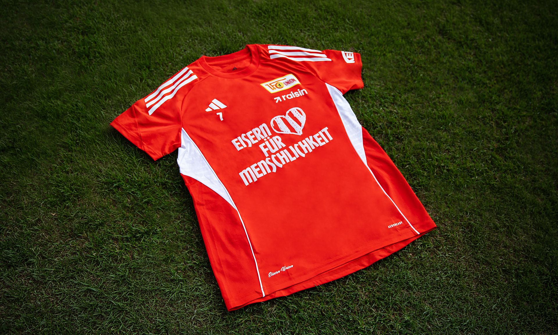 Rotes Fußballtrikot mit weißen Details und dem Schriftzug "EISERN FÜR MENSCHLICHKEIT" auf der Vorderseite, liegend auf grünem Rasen.
