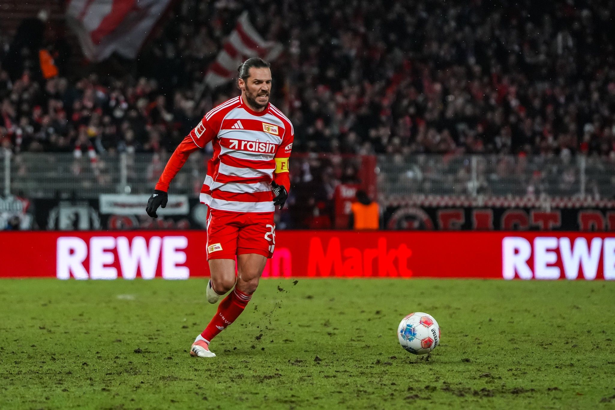 Ein Fußballspieler in rotem Trikot mit der Nummer 23 sprintet über das Feld, während Fans im Hintergrund jubeln.
