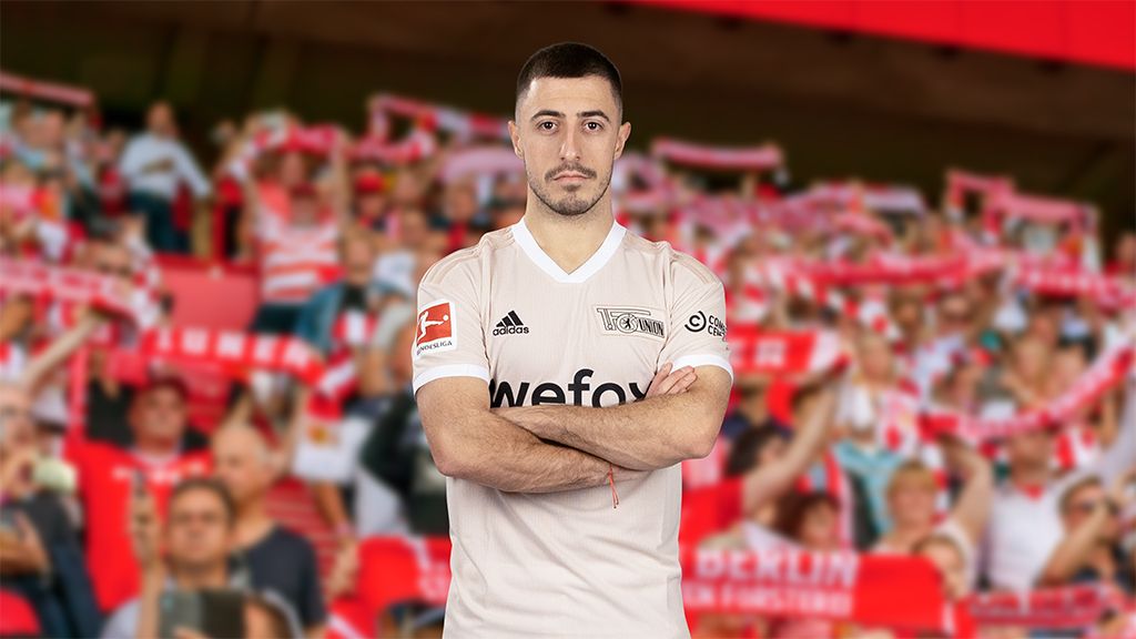Ein Fußballspieler in einem beigen Trikot steht vor einer Menge von Fans, die Schals halten und jubeln.