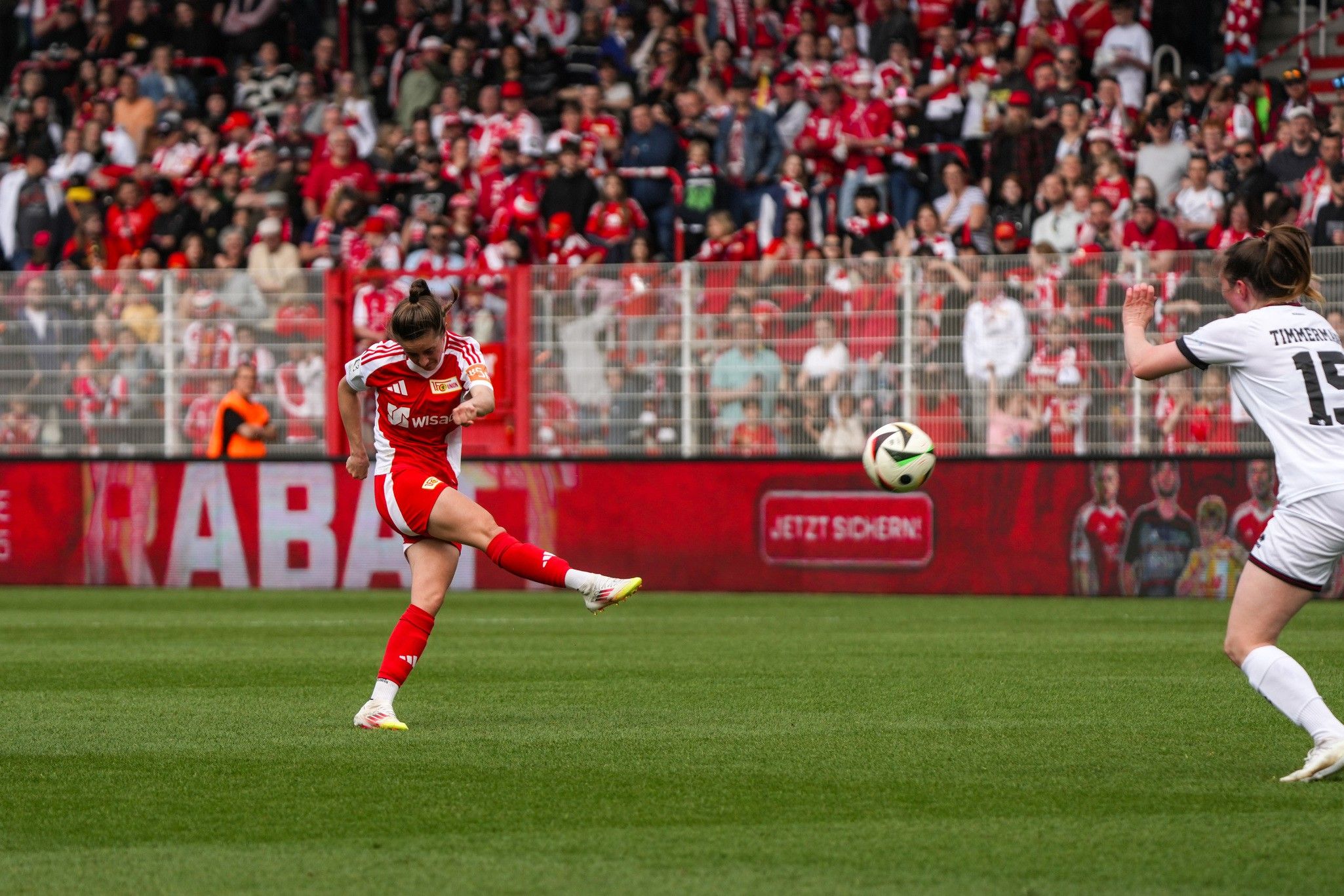 Eine Fußballspielerin in rotem Trikot schießt den Ball während eines Spiels vor jubelnden Fans im Hintergrund.