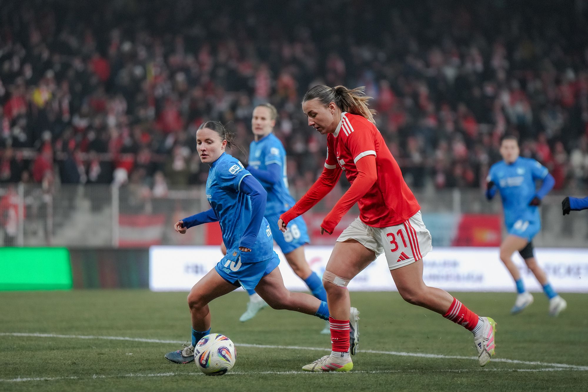 Zwei Fußballspielerinnen in Konkurrenz um den Ball auf einem Sportplatz, im Hintergrund jubelnde Fans.