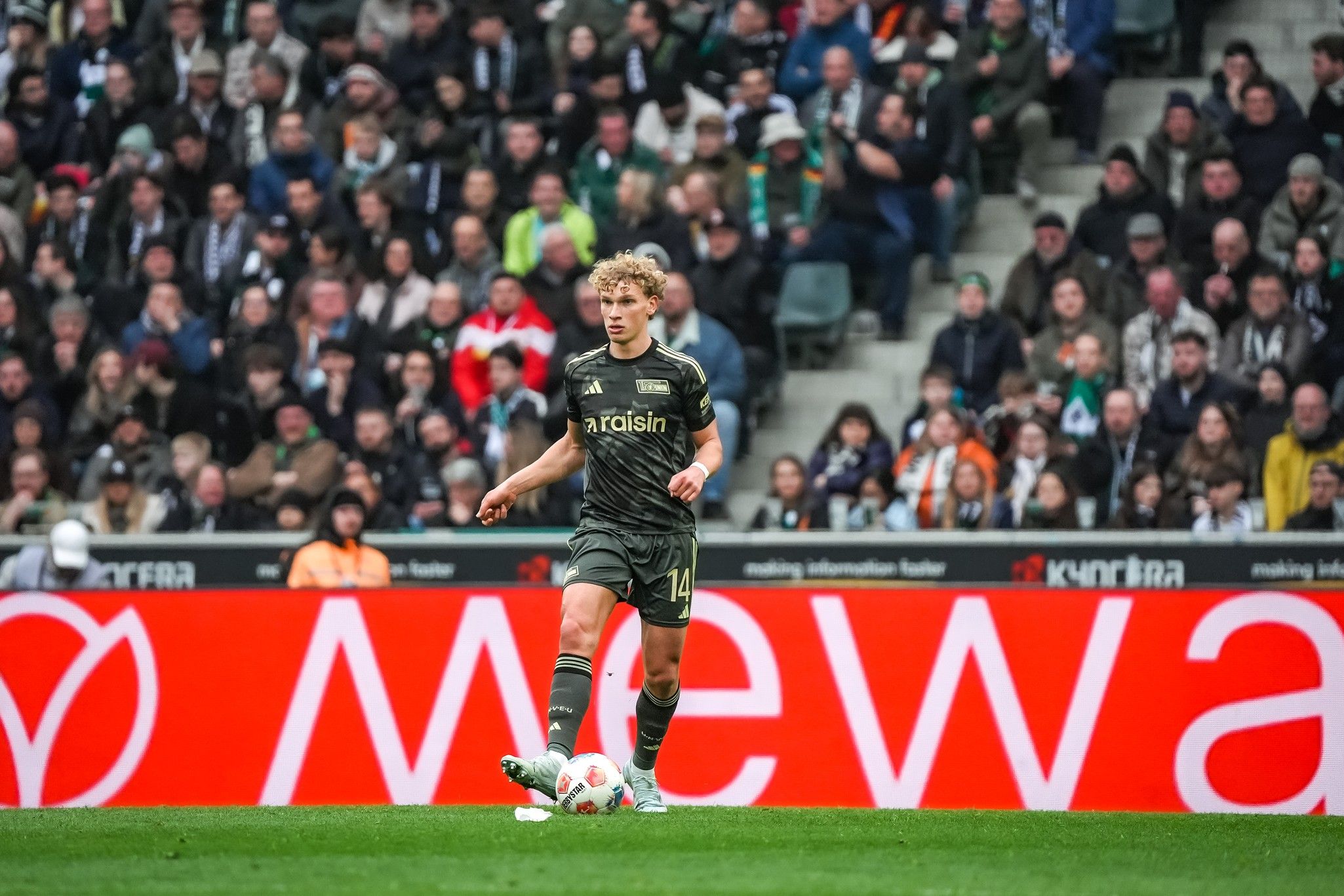 Ein Fußballspieler trägt ein graues Trikot mit der Nummer 14 und spielt vor einer jubelnden Menge im Stadion.