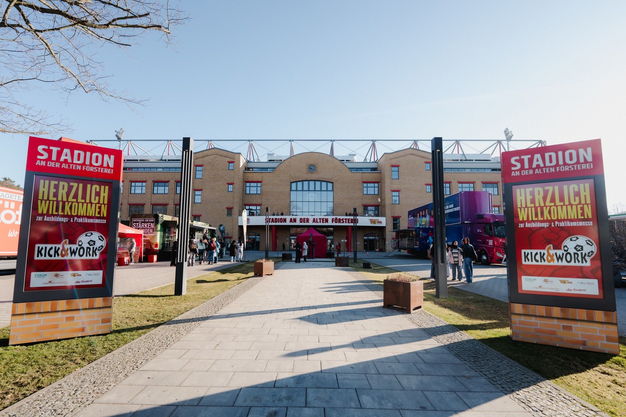 Stadion mit zwei großen Willkommensschildern, umgeben von einem Weg und Gebäuden im Hintergrund.