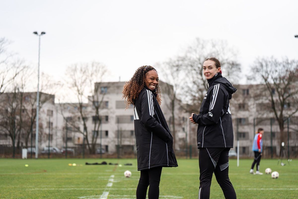 Zwei Fußballspielerinnen stehen auf einem Trainingsfeld, lächeln und tragen schwarze Trainingsjacken. Im Hintergrund sind Wohngebäude sichtbar.