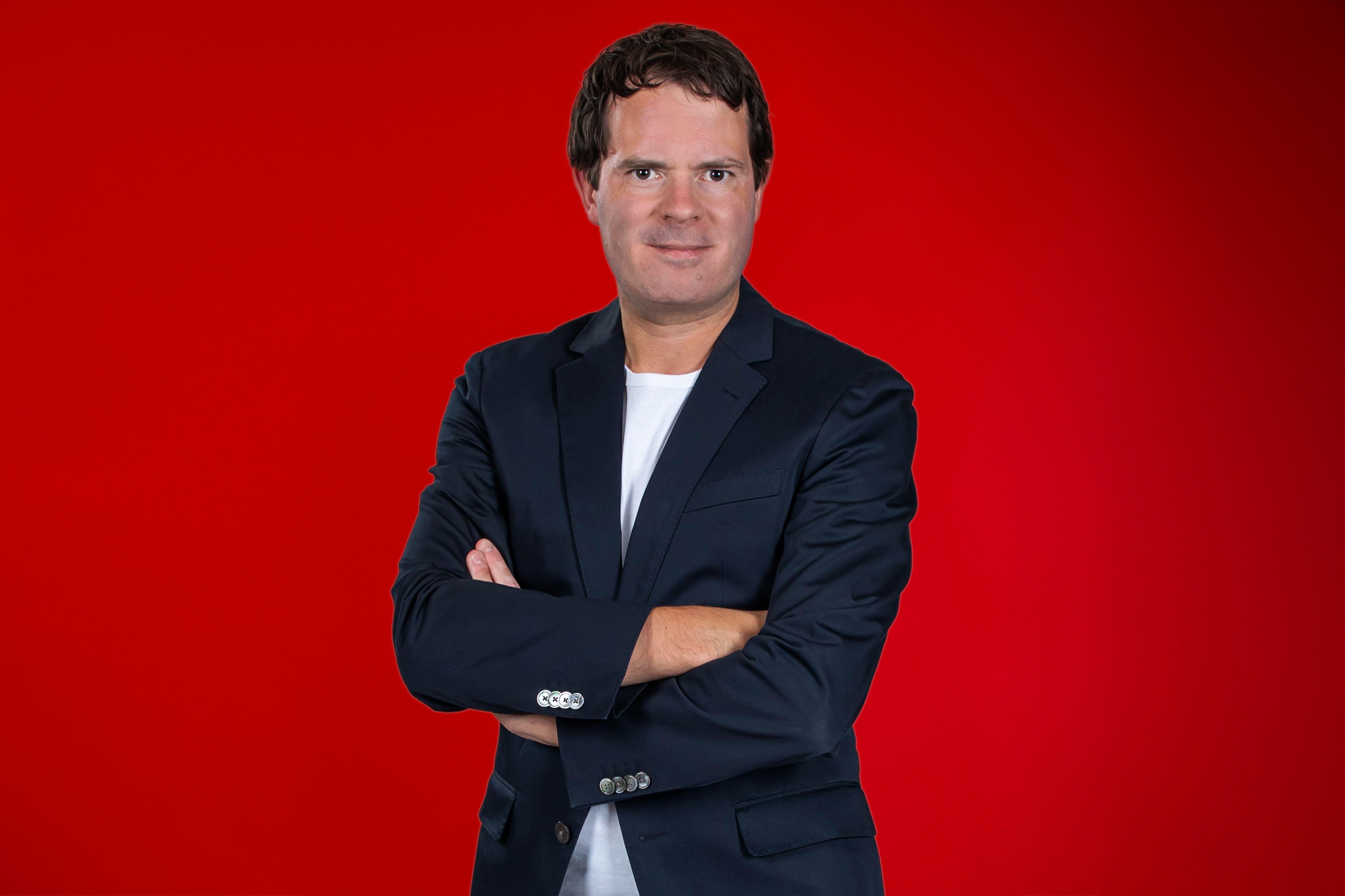 Ein Mann in einem dunkelblauen Blazer und weißem T-Shirt steht mit verschränkten Armen vor einem roten Hintergrund.