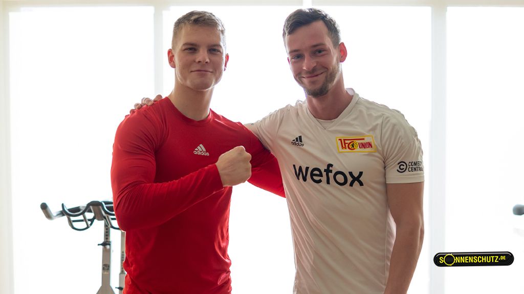Two men smile together in a gym, one in a red shirt and the other in a sports jersey.