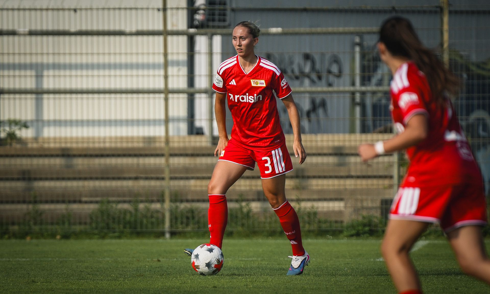 Eine Fußballspielerin in rotem Trikot dribbelt den Ball auf einem Rasenfeld, während eine andere Spielerin im Hintergrund steht.