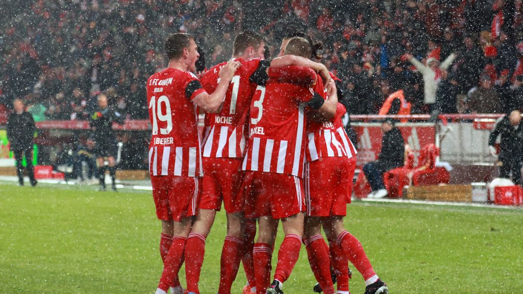 Players in red jerseys embrace joyfully on a soccer field while it snows.