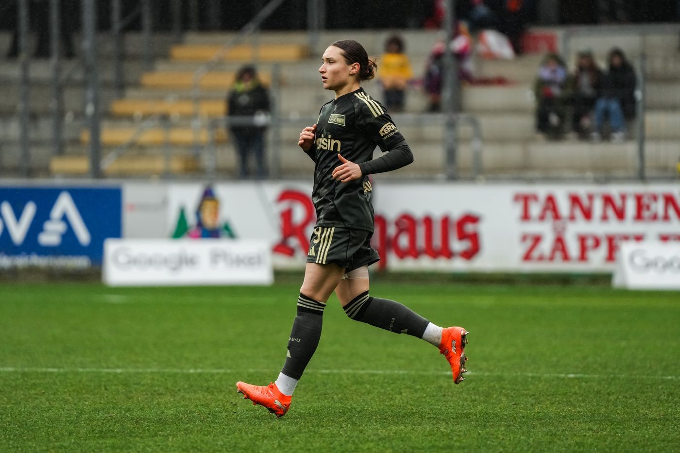 Spieler in schwarzer Fußballausrüstung joggt auf dem Spielfeld, im Hintergrund sind Zuschauer und Werbebanner sichtbar.