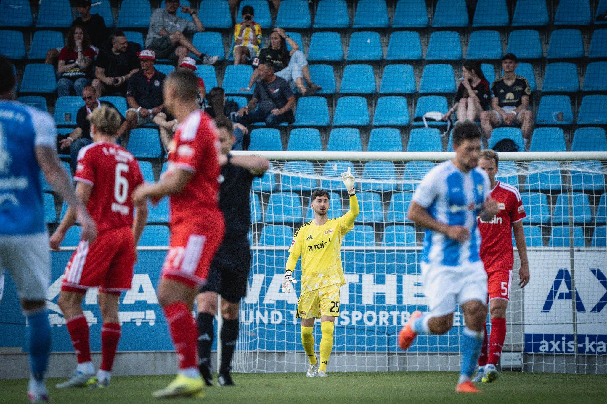 Fußballspiel mit Spielern in Rot und Blau, Torwart in Gelb steht im Tor, Fans im Hintergrund.