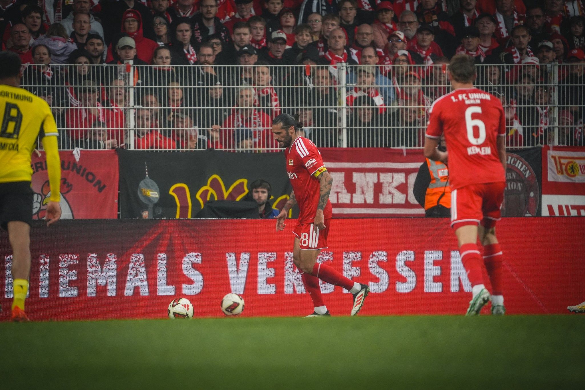 Fußballspiel mit Spielern in roten Trikots und Fans im Hintergrund; ein Spieler bereitet sich auf einen Freistoß vor.