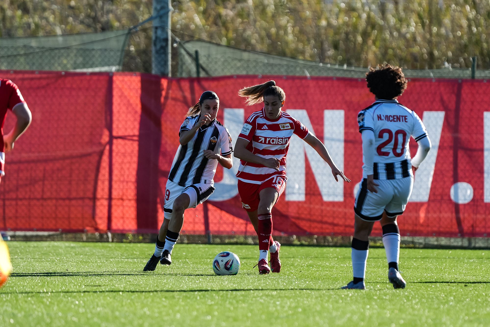 Zwei Soccer-Spielerinnen im Wettlauf um den Ball, während eine Spielerin verfolgt wird, im Hintergrund ein rotes Mannschaftslogo.