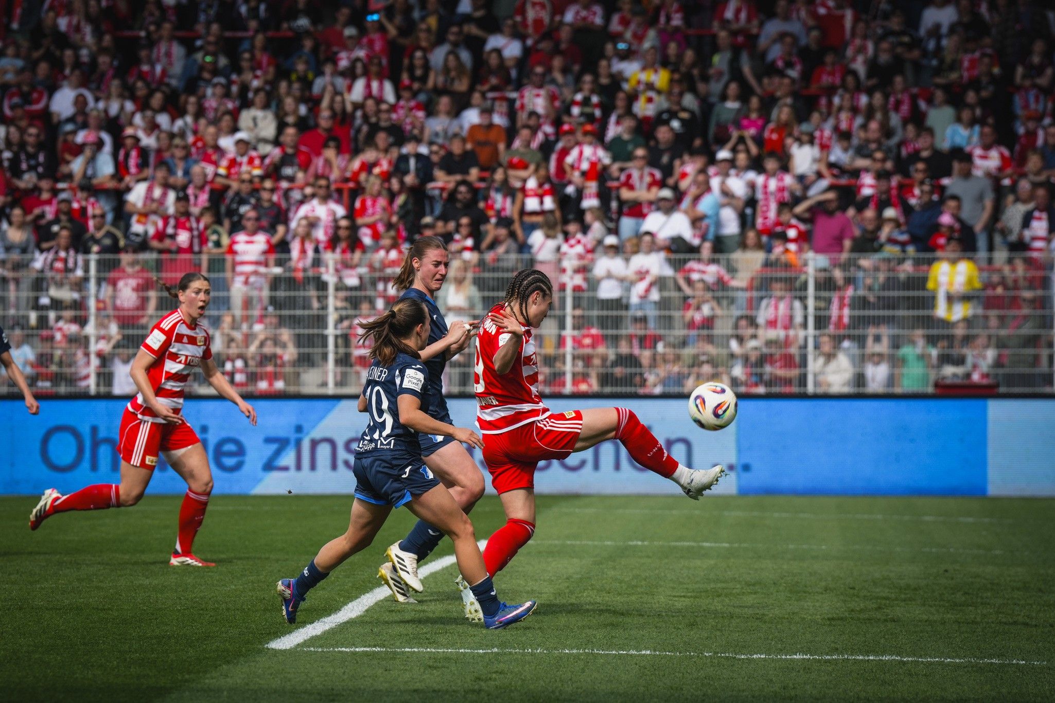 Spielszene im Fußball mit zwei Spielerinnen, die um den Ball kämpfen, im Hintergrund eine jubelnde Menge.