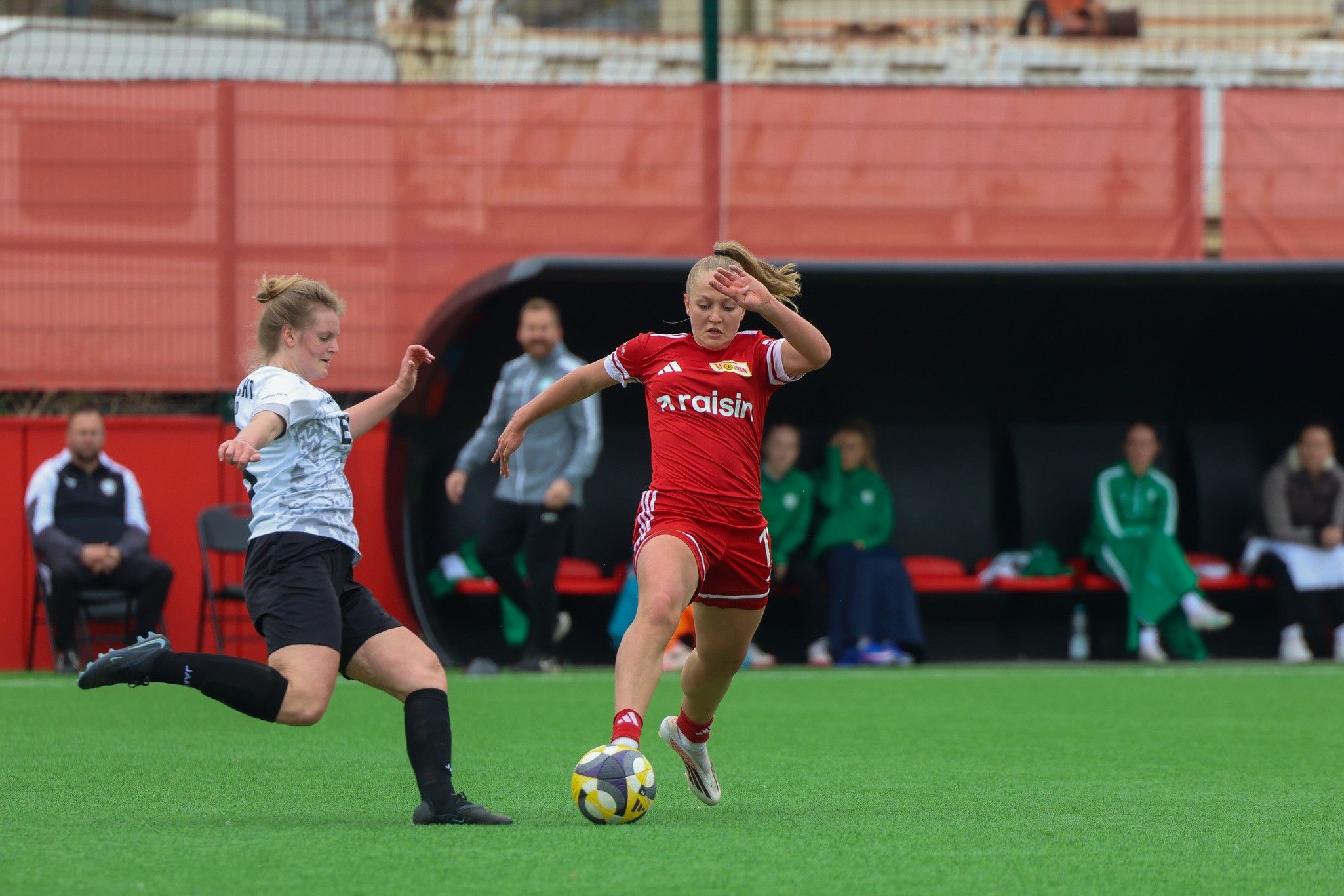 Eine Spielerin in rotem Trikot läuft mit dem Ball, während eine Gegnerin in Schwarz versucht, sie zu stoppen.