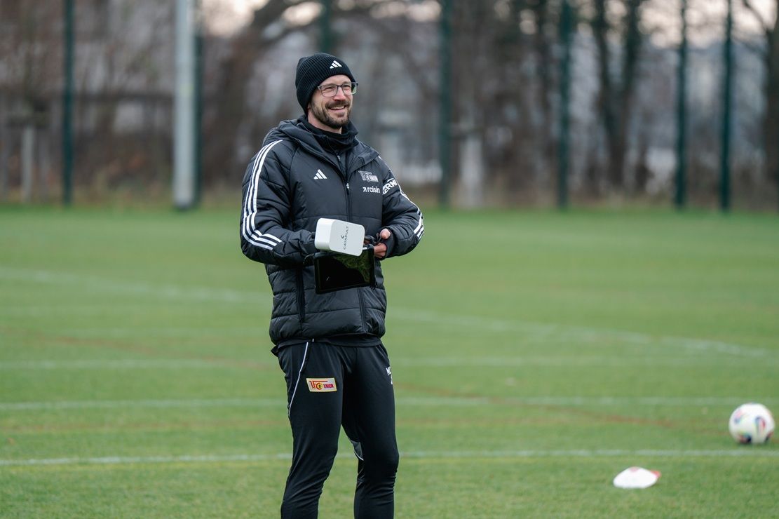 Ein lächelnder Trainer in sportlicher Kleidung steht auf einem Fußballfeld und hält ein Tablet in der Hand.