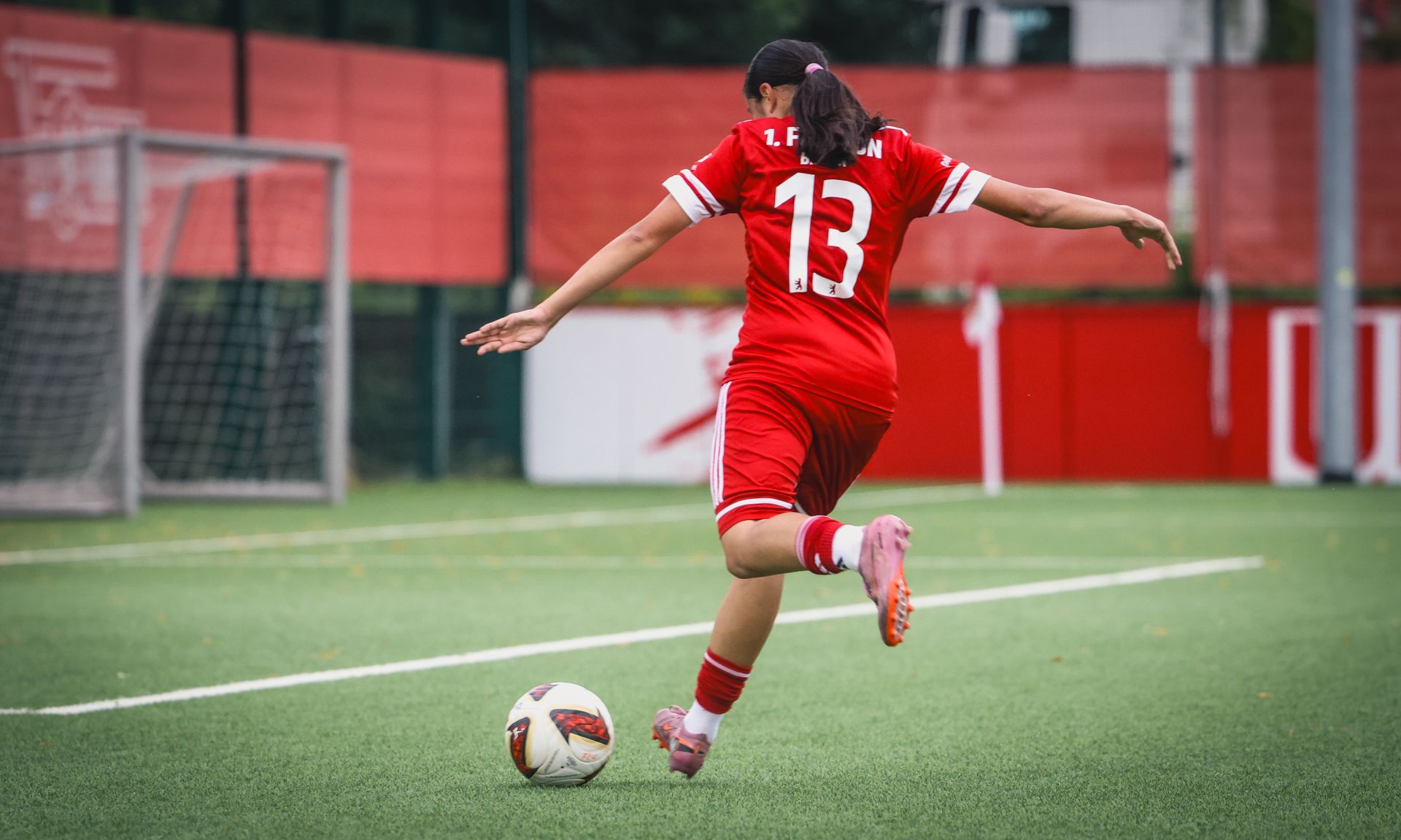 Eine Spielerin in rotem Trikot mit der Nummer 13 tritt einen Fußball auf einem Kunstrasenplatz.
