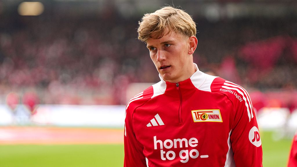 Ein junger Fußballspieler in rotem Trainingsanzug steht auf dem Spielfeld und schaut konzentriert.