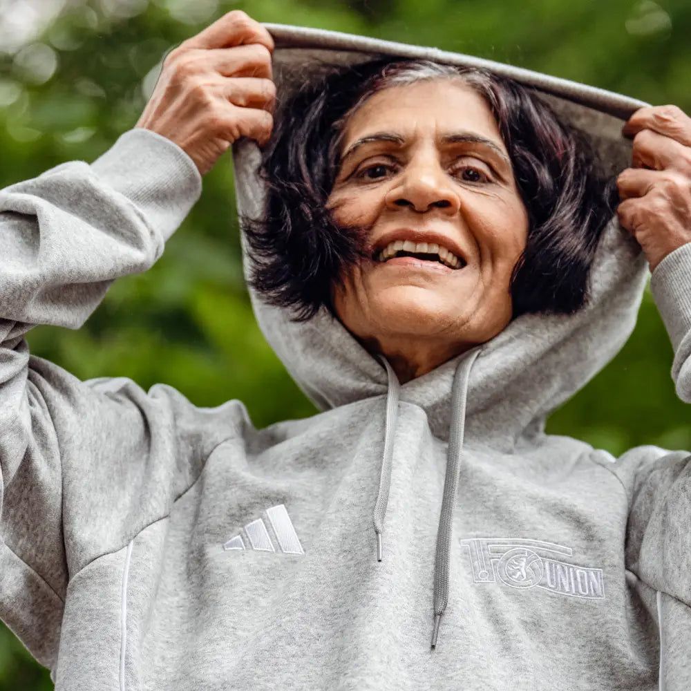 Eine alte Frau hat schwarzes Haar. Sie trägt einen grauen Hoodie. Sie lächelt und zieht den Hoodie über ihren Kopf.