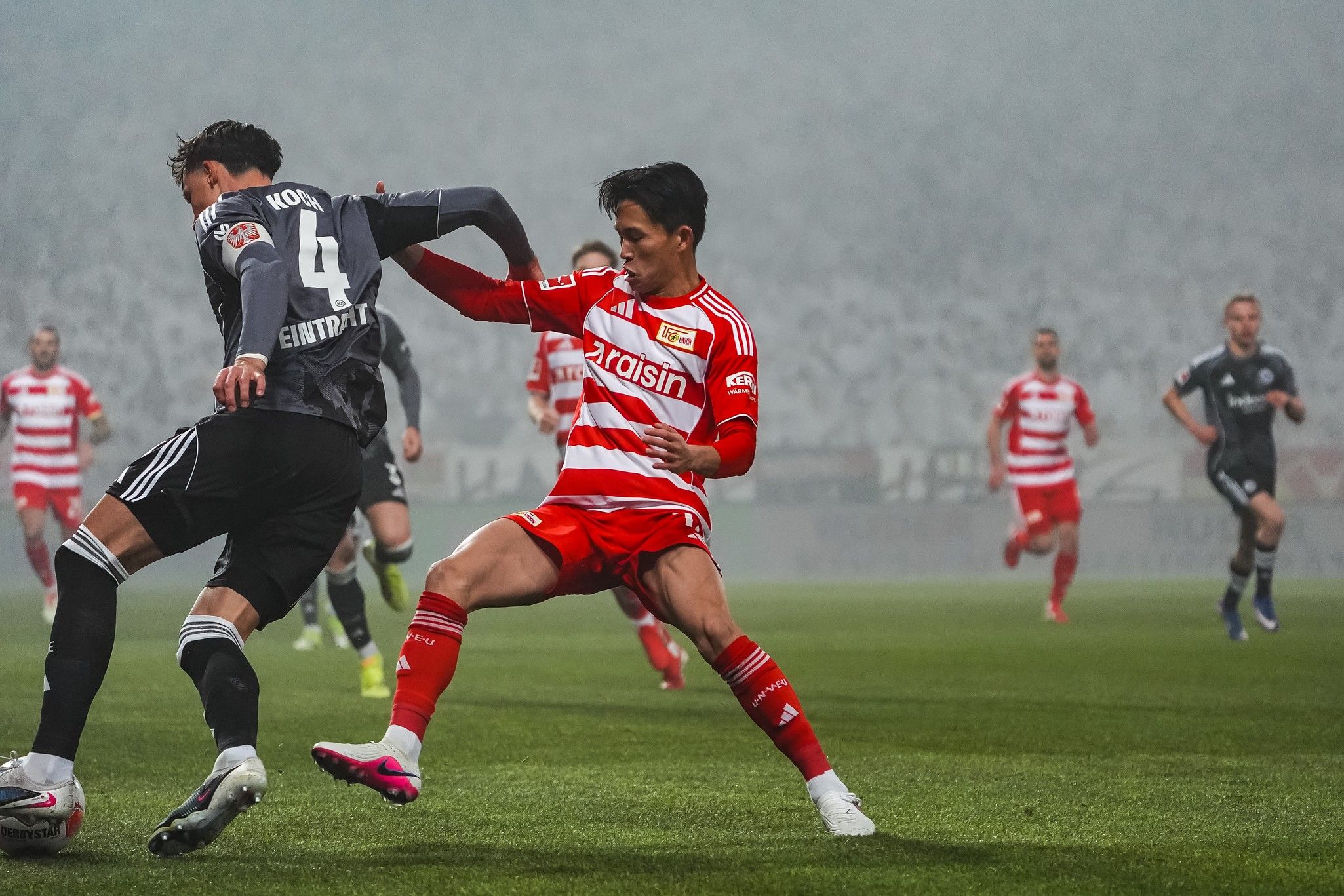 Spielszene im Fußball mit zwei Spielern in Aktion, einer in rotem Trikot und der andere in grauem. Nebel umgibt das Spielfeld.