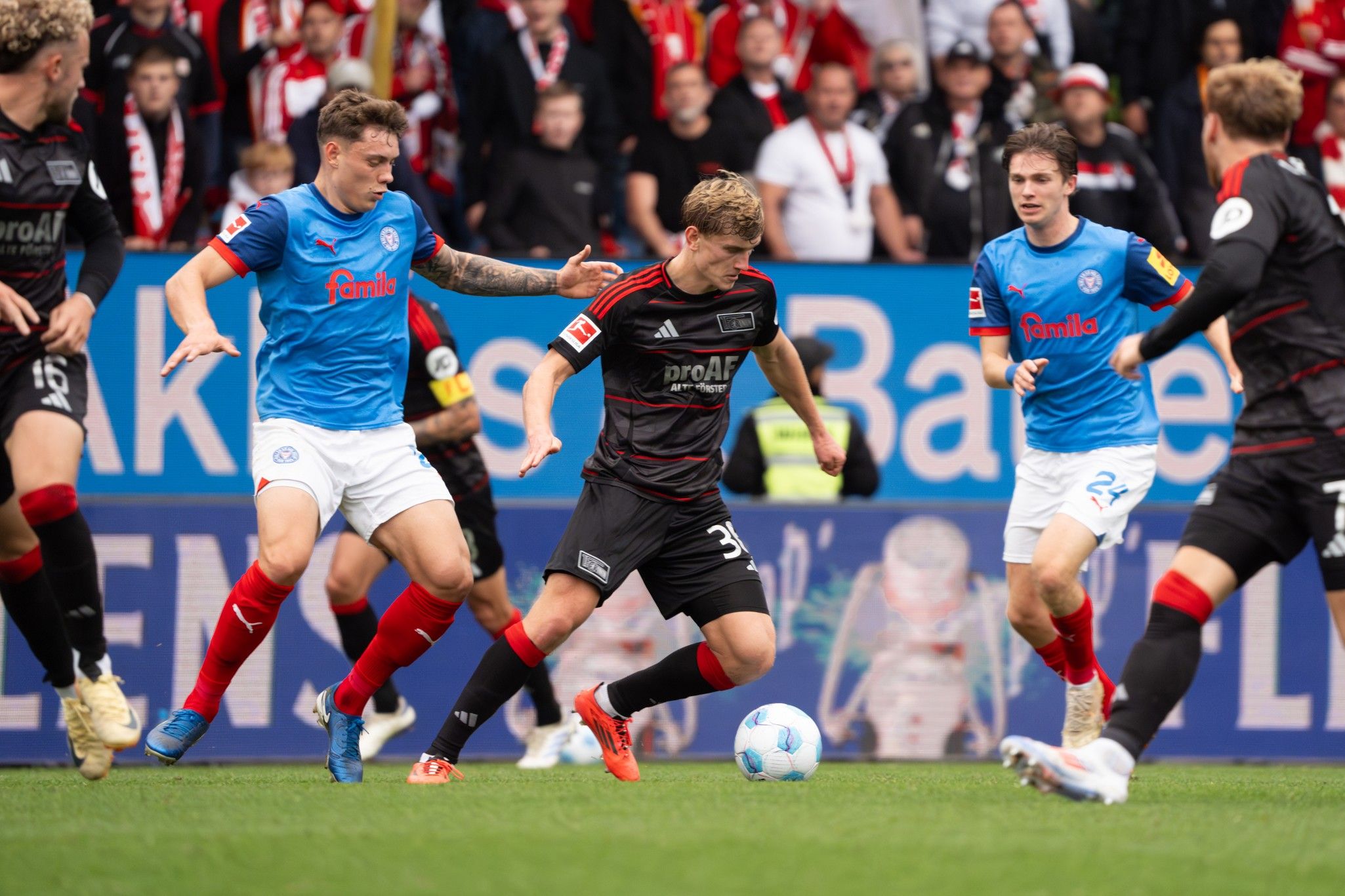 Spielszene aus einem Fußballmatch mit Spielern in schwarz und blau, die um den Ball kämpfen, vor einer jubelnden Menge.
