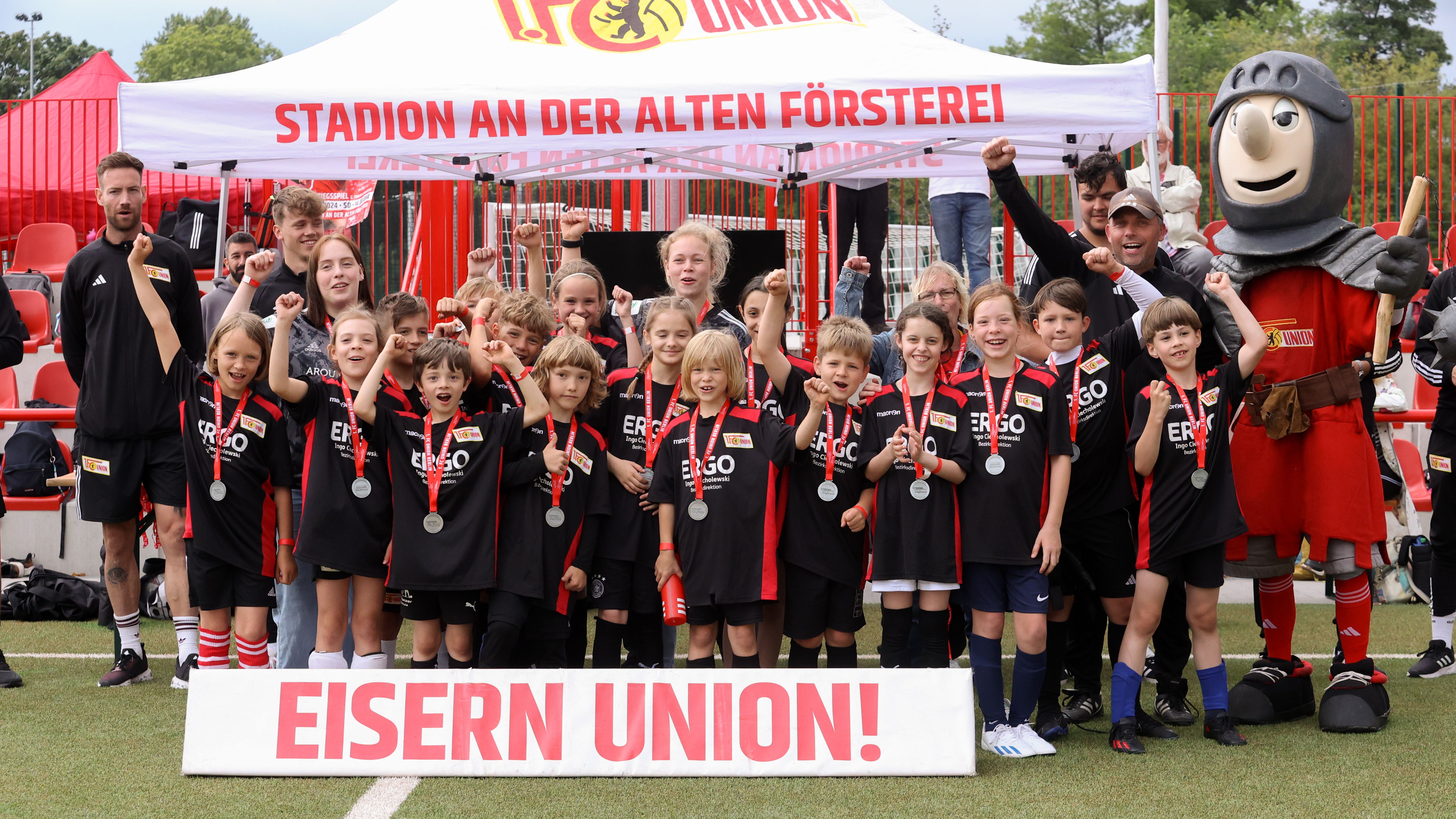 Gruppenfoto von Kindern in Fußballtrikots mit Medaillen, die vor einem Banner mit der Aufschrift "EISERN UNION!" stehen.