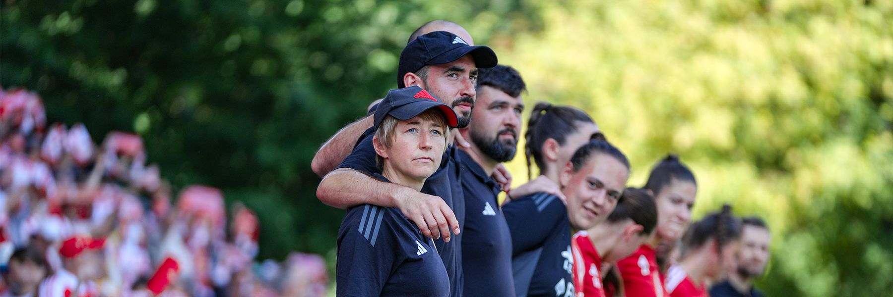A group of athletes stands together, some wearing black clothing and caps, with spectators visible in the background.