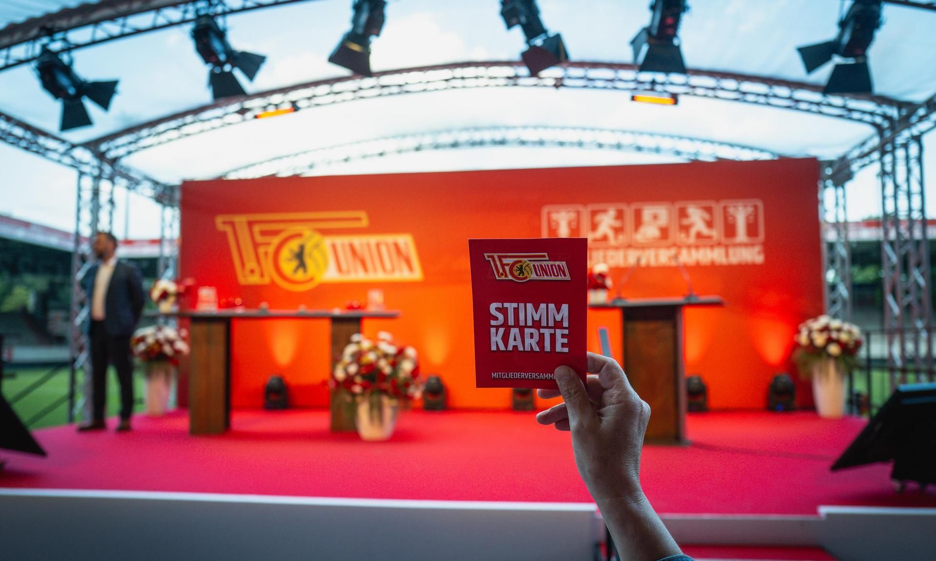 Eine Hand hält eine rote Stimmkarte vor einer Bühne mit dekorativen Blumenarrangements und Beleuchtung im Hintergrund.
