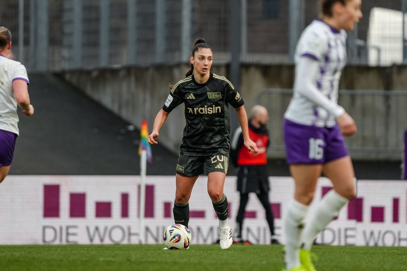Spielerin in dunkler Sportkleidung dribbelt den Ball auf dem Spielfeld, während andere Spieler im Hintergrund agieren.