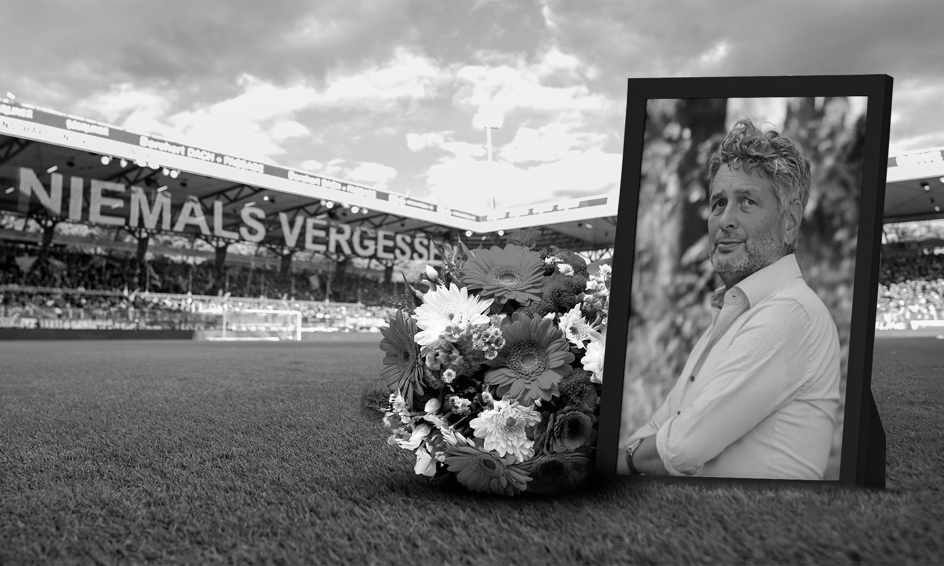 Ein Blumenkranz und ein gerahmtes Porträt eines Mannes vor einem Stadion mit der Aufschrift „NIEMALS VERGESSEN“ im Hintergrund.