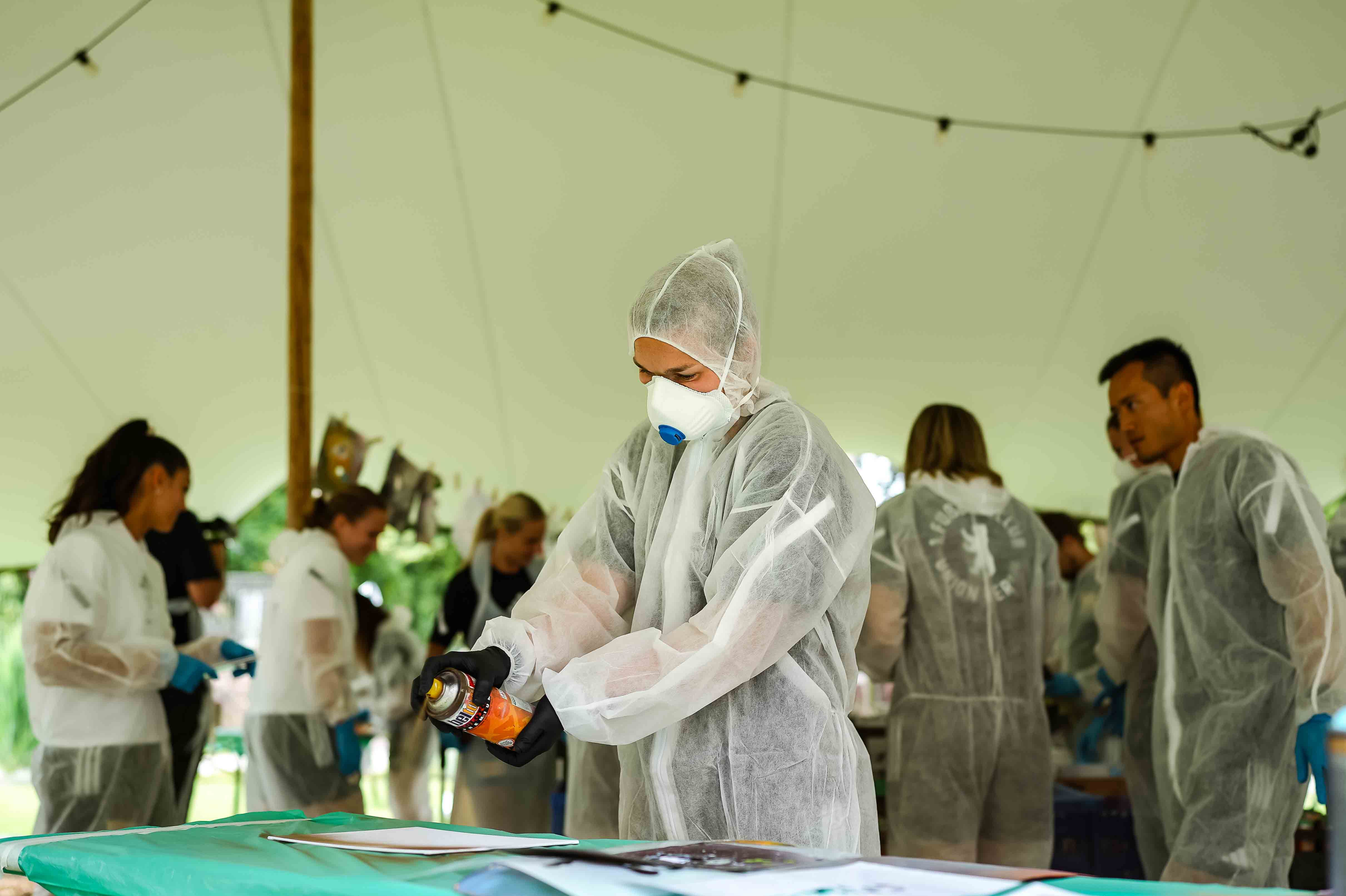 Person in Schutzanzug und Maske arbeitet an einem Tisch unter einem Zelt, umgeben von weiteren Personen in ähnlicher Ausrüstung.