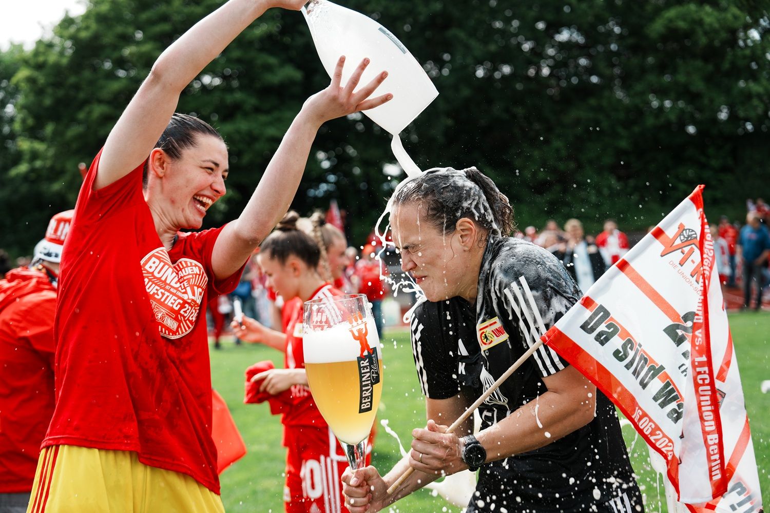 Zwei Frauen feiern mit Bier und einer Fahne auf einem Fußballfeld, während Wasser über eine von ihnen geschüttet wird.