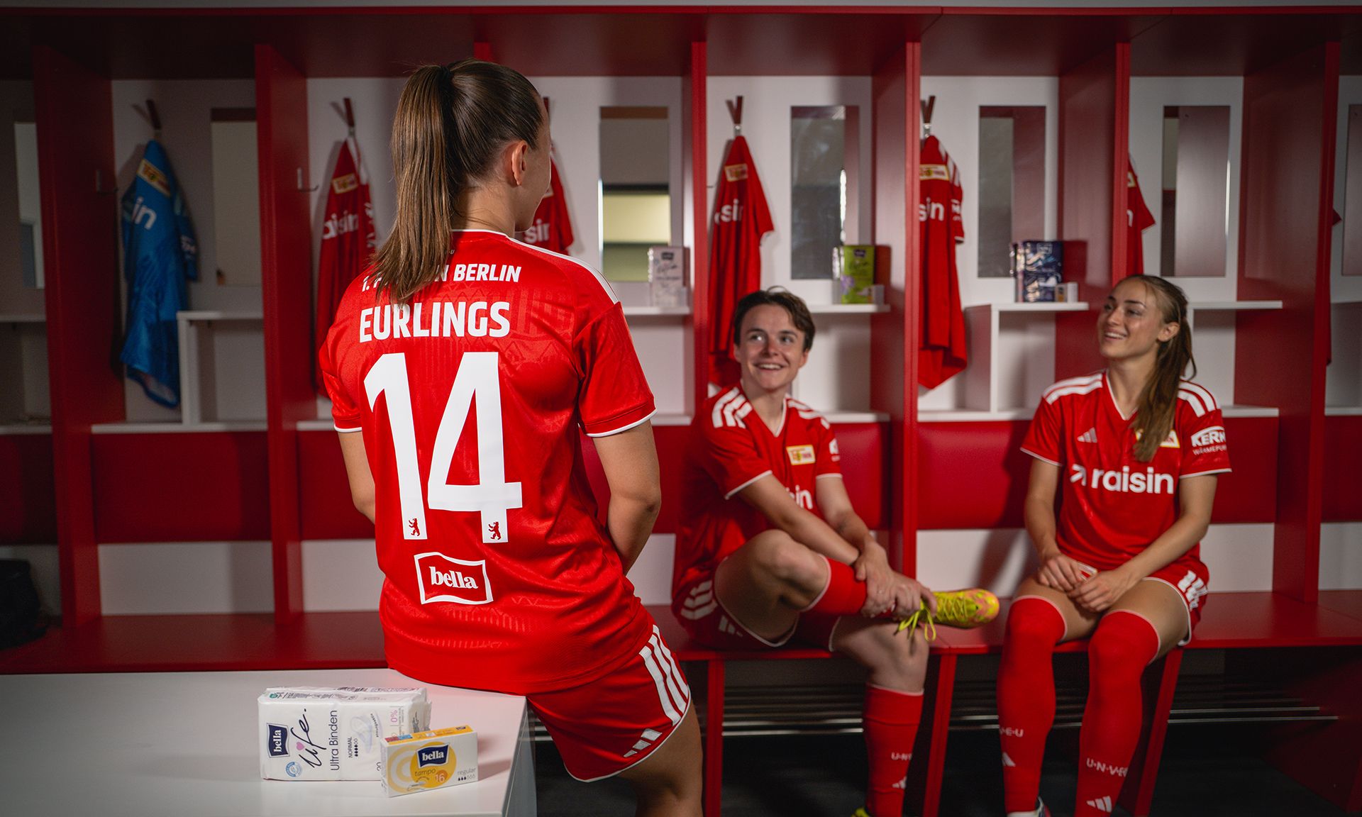 Drei Fußballspielerinnen im roten Trikot sitzen in einer Umkleidekabine, während eine von ihnen in die Kamera schaut.