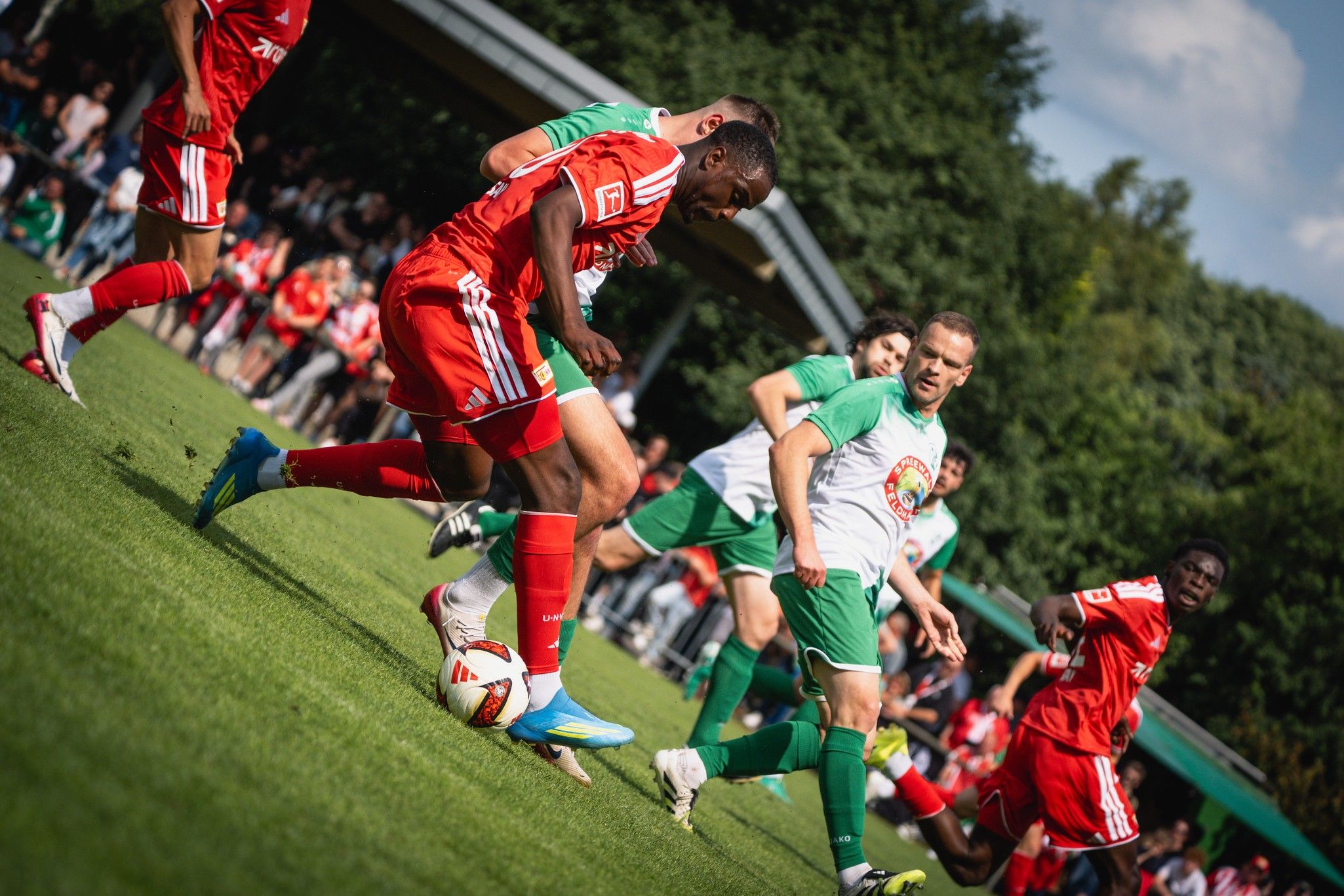 Ein Fußballspiel mit Spielern in roten und grünen Trikots auf einem Rasenplatz, umgeben von Zuschauern.