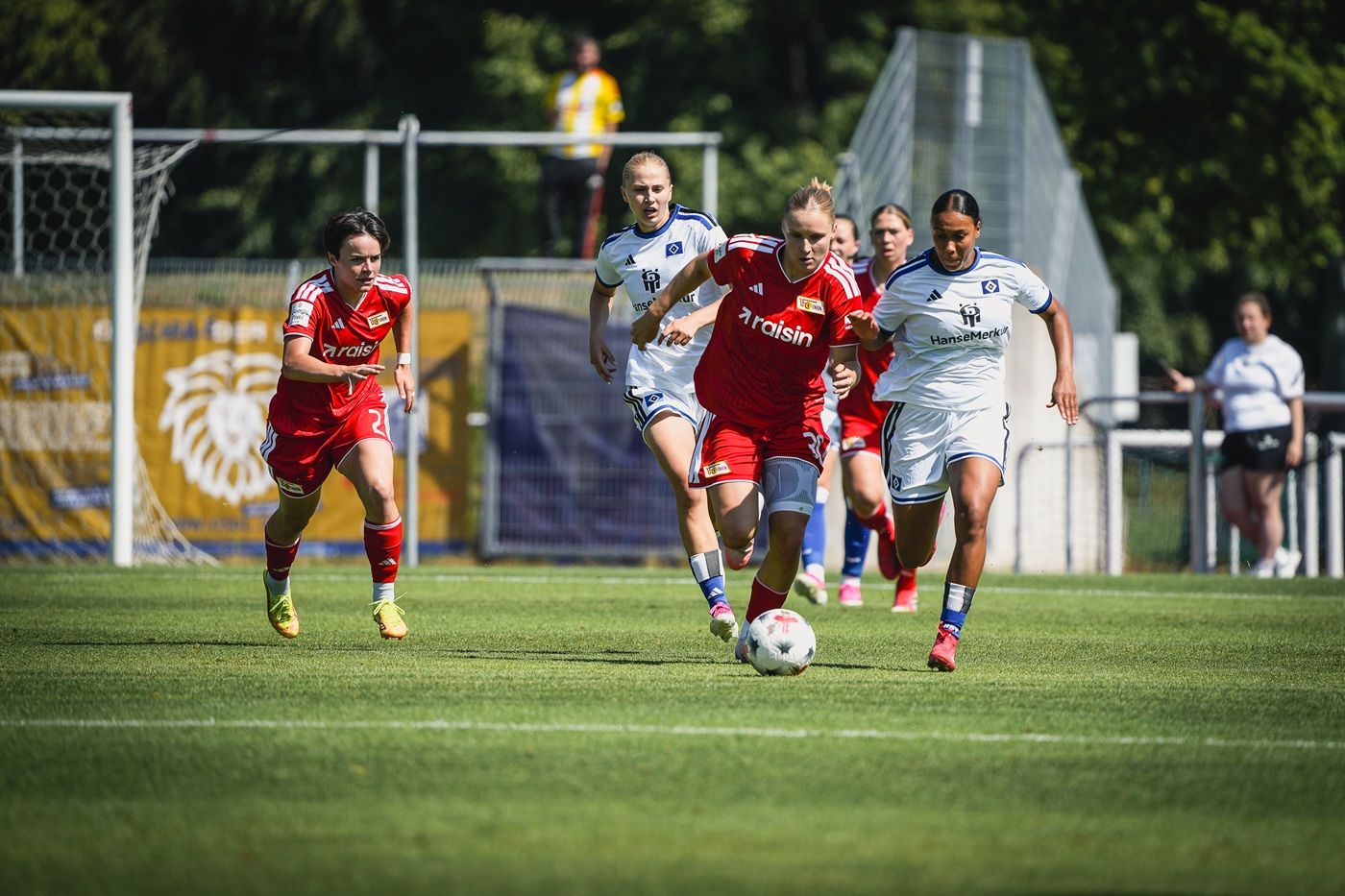 Eine Spielerin in roter Kleidung sprintet mit dem Ball, während sie von zwei Gegnerinnen in Weiß verfolgt wird.