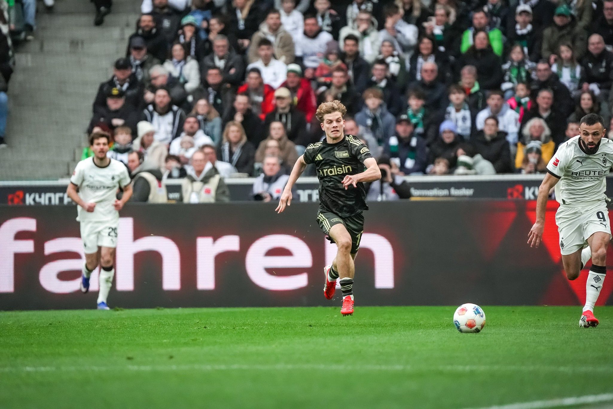 Spielszene im Fußball mit einem Spieler in Schwarz, der dem Ball entgegenläuft, während Zuschauer im Hintergrund zu sehen sind.