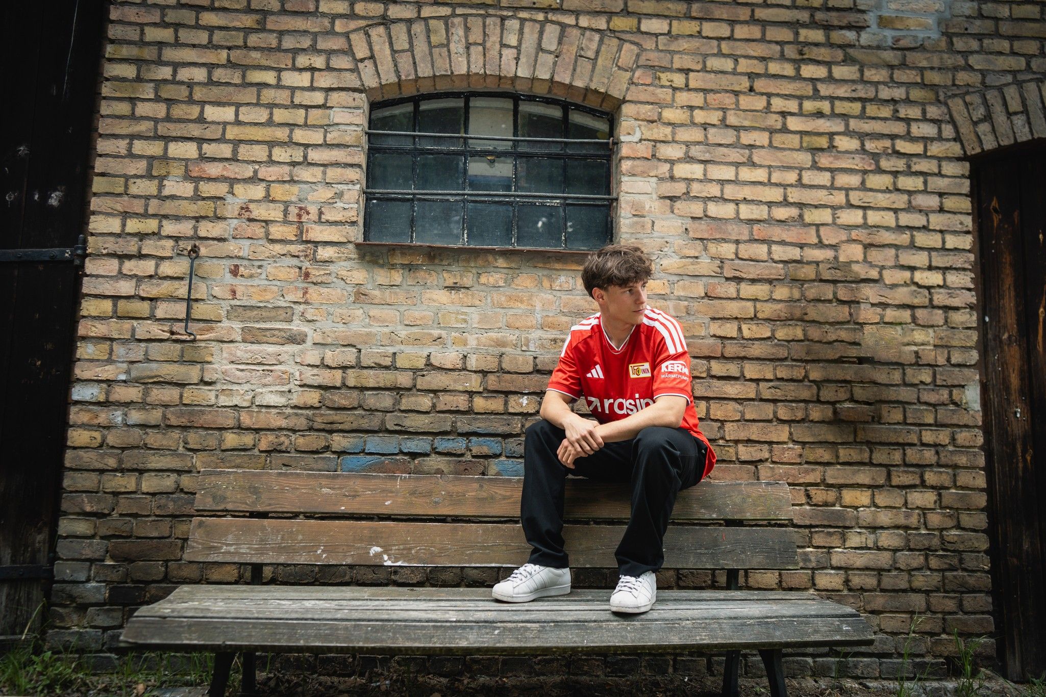Ein junger Mann in einem roten Fußballtrikot sitzt auf einer Bank vor einer Backsteinmauer.